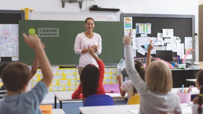 Rear view of school kids raising hand while they are sitting on their chair with their teacher in front of them against a greenboard in background